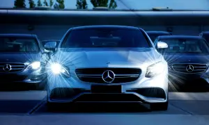 Biała hybryda plug-in Mercedes-Benz z włączonymi światłami, otoczona innymi ciemnymi samochodami tej marki.