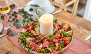 Antipasti selber machen: Eine festliche Platte mit Wurst, Käse, Brot, Oliven und Tomaten, umgeben von grünen Zweigen, mit einer Kerze in der Mitte.