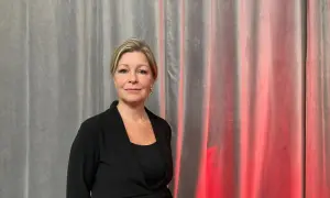 Johanna Fager Wettergren, en kvinna med blont hår och blå ögon, ler mot kameran framför en grå ridå med röd belysning.