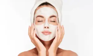 Une femme avec une serviette sur la tête applique un masque blanc sur son visage, espérant atténuer les marques de masque cicatrice acné.