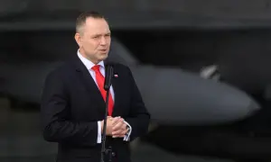 Mężczyzna w garniturze przemawia przy mikrofonie, w tle samolot. Mówi o reparacje wojenne od niemiec.