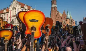 Tłum ludzi z gitarami wzniesionymi ku górze, świętujący gitarowy rekord świata na tle zabytkowej architektury.