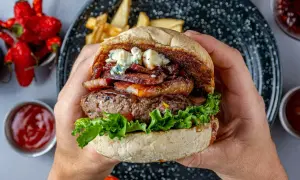 Ręce trzymają soczystego burgera z serem pleśniowym, boczkiem i sałatą. Obok frytki i sosy.