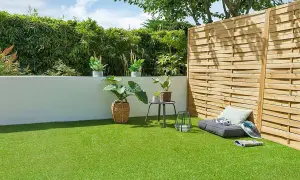Un jardín moderno con césped artificial verde vibrante, rodeado de plantas y una valla de madera. Un rincón acogedor con cojines invita al descanso.
