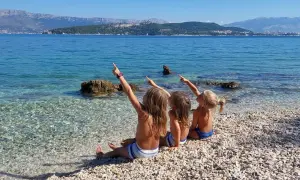 Trzy dziewczynki siedzą na kamienistej plaży, wskazując palcami na morze i góry. Mówią sobie "Dzień dobry po chorwacku" w słoneczny dzień.