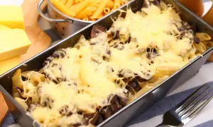 Pyszna zapiekanka z pieczarkami i makaronem penne, zapieczona pod złotym serem. Obok surowy makaron i kawałki sera.