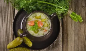 Aromaticzna zupa ogórkowa na żeberkach, podana w brązowej miseczce, ozdobiona koperkiem. Obok leżą chrupiące ogórki kiszone i pęczek świeżego koperku.