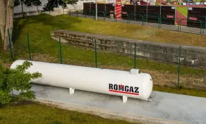 Biały zbiornik gazu ROMGAZ stoi na betonowej podstawie. Widać ogrodzenie i trawę, a w tle trybuny stadionu. Odległość gazociągu od ogrodzenia jest widoczna.
