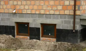 Prace przy izolacji fundamentów w starym domu. Dwa okna piwniczne, czarna izolacja przeciwwilgociowa i nowe mury.