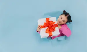 Uśmiechnięta dziewczynka z prezentem w rękach, wyłaniająca się z błękitnej dziury w papierze. Idealny obrazek na dzień dziecka!