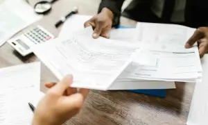 Dwie osoby wymieniają się dokumentami na biurku z kalkulatorem i lupą. Czekamy na wynik analizy.