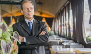 Uomo d'affari con braccia conserte, un chief people officer che guarda lontano, con piante verdi in primo piano e un ristorante sullo sfondo.
