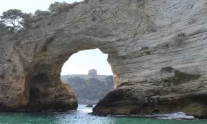 Arco naturale di roccia che incornicia una torre sulla collina, vista dalla baia San Felice Vieste. Acque turchesi e vegetazione mediterranea.