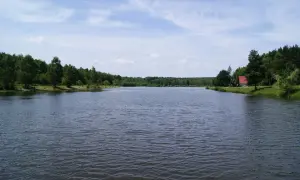 Spokojna tafla jeziora, otoczona zielenią lasów, z małym czerwonym domkiem na brzegu. Wesoła fala Mysłowice zaprasza do odpoczynku.