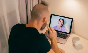 Mężczyzna odbywa konsultację psychologiczną online. Na ekranie laptopa widoczna uśmiechnięta kobieta machająca ręką.