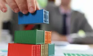 Dłoń układa niebieski kontener na stosie kolorowych modeli. Analiza kosztów transportu i potencjalnego cła.