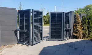 Otwarta brama łamana, szara, z lamelkami, z automatyką, na tle zieleni i nieba.