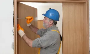 Hombre con casco azul y guantes naranjas ajusta una puerta de contrachapado con un destornillador.