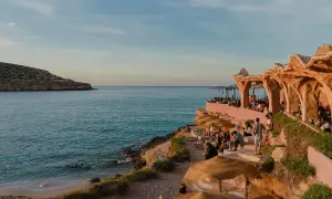 Menschen genießen den Sonnenuntergang am Strand von Ibiza, in der Nähe eines einzigartigen ashram-ähnlichen Gebäudes am Meer.