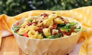 Ein köstlicher tortellini salat mediterran mit sonnengetrockneten Tomaten und Nüssen, serviert in einer weißen Schüssel auf einem Holztisch.