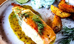 Gebratener Lachs mit Kräuterdressing, Kartoffelspalten und einem Klecks Gurkensalat. Ein warmes Gericht, das an Gurken Rezepte warm erinnert.