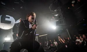 The Hives setlist is being performed by a charismatic frontman in a black suit, belting into a microphone on a smoky stage. The crowd is visible, enjoying the energetic show.