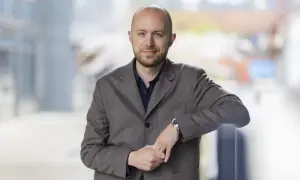 Viktor Barth Kron, en man med kortklippt hår och skägg, bär en brun kavaj och en mörkblå skjorta. Han lutar sig mot ett räcke utomhus.