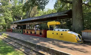 Die gelbe Lokomotive der Schlossbahn Karlsruhe steht bereit. Fahrgäste sitzen in den roten und grünen Waggons unter einem schattigen Dach.