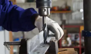 Ręka w rękawicy obsługuje wiertarkę stołową, wiercąc w metalowym profilu. Widać części do wiertarki stołowej i wióry metalowe.