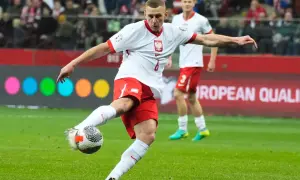 Piłkarz w białej koszulce z orłem i czerwonych spodenkach kopie piłkę na boisku.