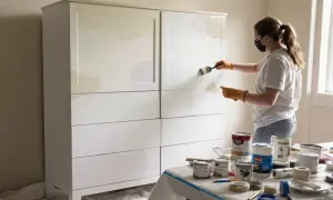 Kobieta maluje białe mdf meble na jasny kolor. Na stole stoją puszki z farbą i taśmy malarskie.