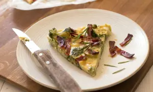 Une part de quiche sans pâte aux brocolis et lardons, garnie de romarin, sur une assiette avec un couteau.