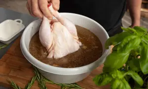Ein rohes Hähnchen wird in eine Schüssel mit einer würzigen Flüssigkeit getaucht, um Fleisch in Salzlake einzulegen. Rosmarin und Basilikum liegen daneben.