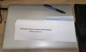 Na biurku leży laptop, na nim dokument z napisem "Rozwiązanie umowy o pracę bez wypowiedzenia (zwolnienie dyscyplinarne)". Obok długopis i myszka.