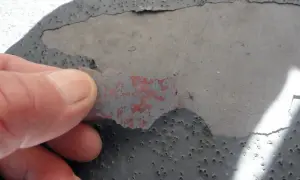Dedo retirando capa desprendida de pintura gris sobre hormigón con picaduras, indicando necesidad de reparación de hormigón.