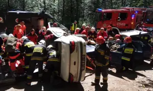 Akcja ratunkowa po wypadku. Strażacy KSRG zabezpieczają teren i wyciągają poszkodowanych z przewróconego auta. W tle wozy strażackie i karetka.