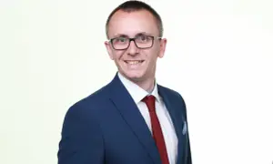 Mężczyzna w garniturze i okularach, uśmiecha się na białym tle.