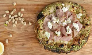 Pizza mit Mortadella, Pistazien und cremigem Käse, daneben einige Pistazien und eine Zitronenspalte.