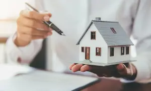 Dłoń trzymająca model domu i długopis, sugerujące, co jest potrzebne do ubezpieczenia domu.