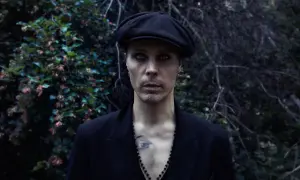 Ville Valo w czarnym kaszkiecie i marynarce, z intensywnym makijażem oczu, stoi na tle ciemnej, leśnej roślinności.