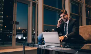 Uśmiechnięty mężczyzna w garniturze rozmawia przez telefon, pracując przy laptopie. To jego rozliczenie delegacji krajowej.