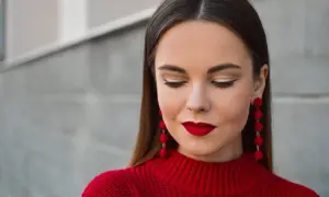 Une femme arbore un beau maquillage Noël, avec des lèvres rouge vif et des yeux dorés, portant un pull rouge et des boucles d'oreilles assorties.