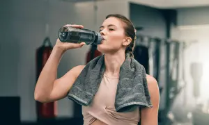 Eine Frau trinkt nach dem Training aus ihrer Trinkflasche. Sie hat ein Handtuch um den Hals.