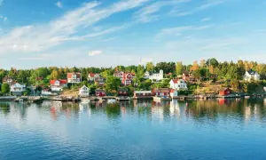 Vackra röda och vita hus längs en grönskande kustlinje, speglande sig i det blå vattnet. En idyllisk vy av sommar i Stockholm.