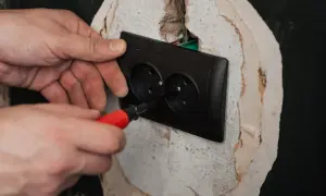 Ręce montują czarne podwójne gniazdko elektryczne. Śrubokręt z czerwoną rączką dokręca śrubę, kończąc podłączenie gniazdka.