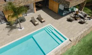 Una piscina azzurra e una moderna pavimentazione esterna in legno creano un'oasi di relax con arredi da esterno e una palma.
