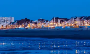 Kvällsljus över strandpromenaden i Dunkirk stad. Husen längs kusten lyser upp mot den mörka himlen, med reflektioner i det lugna vattnet.
