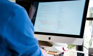 Programista w niebieskiej bluzie pochyla się nad monitorem z kodem.