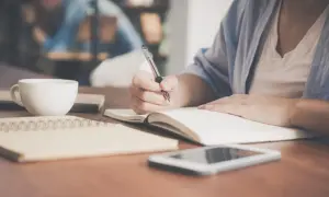 Kobieta pisze w notatniku, obok filiżanka kawy i telefon. Może to być początek ważnej umowy.