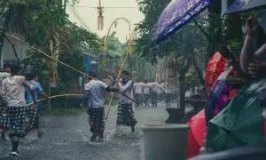 Ulewny deszcz na Bali w marcu. Mężczyźni w tradycyjnych strojach niosą drewniane kije, a widzowie chronią się pod kolorowymi parasolami.